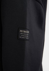 Sweat-shirt noir avec une finition texturée et une étiquette tissée sur la manche portant le texte "PETROLND" ainsi que d'autres détails textuels.