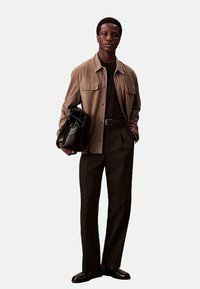 Bruine suède jas over een zwart shirt, gecombineerd met een donkere broek en zwarte schoenen. Het model houdt een zwarte leren tas vast.