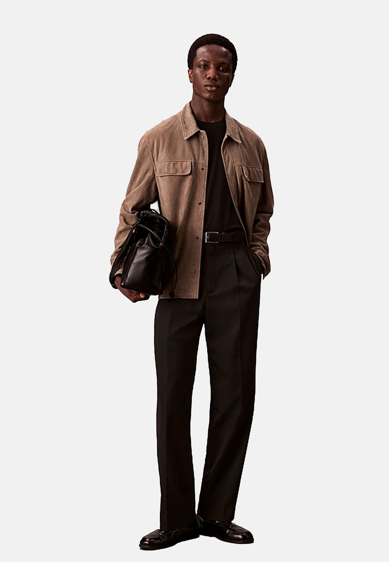 Bruine suède jas over een zwart shirt, gecombineerd met een donkere broek en zwarte schoenen. Het model houdt een zwarte leren tas vast.