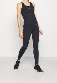 Canottiera sportiva nera con scollatura tonda e design a schiena scoperta. Abbinata a leggings neri con un accento giallo. Indossata con scarpe da ginnastica nere.