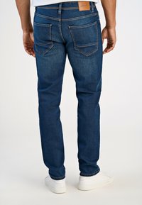 Tummansinisi ä farkkuhousuja, joissa on suora lahkeensuukappale, kaksi takataskua ja nahkapaikka vyötäröllä. Klassinen ompeluviimeistely.