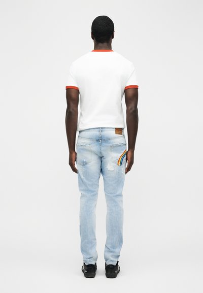 Homme portant un t-shirt blanc avec une bordure orange et un jean bleu clair orné d'un patch arc-en-ciel sur la poche arrière, debout de dos face à la caméra.