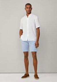 White short-sleeve linen shirt with buttons, light blue shorts, and brown suede slip-on shoes. Model stands against a neutral background.