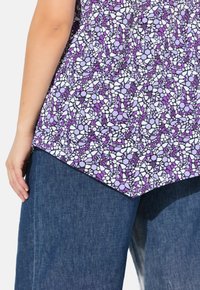 Top viola con motivi irregolari e disegni floreali, abbinato a pantaloni in denim. Il tessuto appare leggero e strutturato.