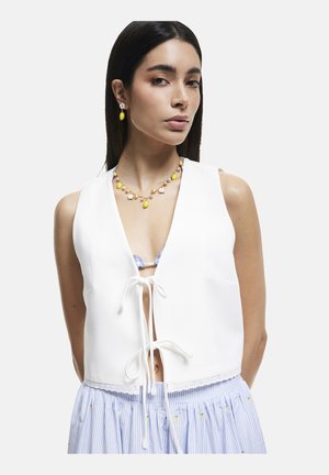 Débardeur court blanc avec un décolleté en V et une fermeture à nouer à l'avant, associé à une jupe rayée bleu clair et blanche avec de petites embellissements.