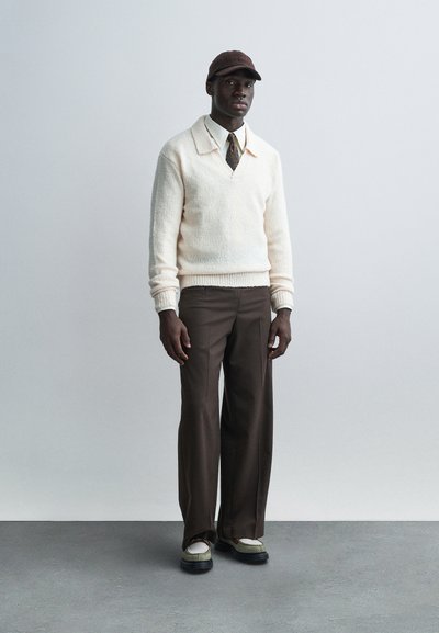 Cream textured knit sweater with a V-neck, paired with brown tailored pants and white sneakers with green accents. Brown cap completes the look.