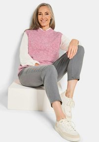 Helles rosa gestricktes ärmelloses Oberteil über einem weißen Langarmhemd, graue cropped Hose und weiße Sneaker; sitzt auf einem weißen Würfel.