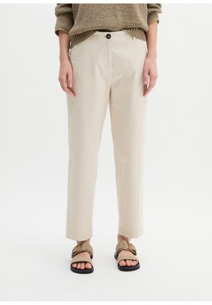 Personne portant un pantalon beige à jambes droites, un pull en tricot marron, et des sandales beige avec des boucles dorées sur un fond blanc.