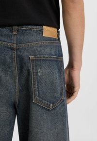 Donkerblauwe denim jeans met oranje stiksels, een leren achterpatch met de oplage van "Hugo," en een traditionele vijf-pocket ontwerp.