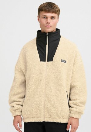 Beige Fleece-Jacke mit einem gesteppte schwarzen Stehkragen, Front-Reißverschluss und Seitentaschen; hat eine strukturierte Oberfläche und eine lässige Passform.