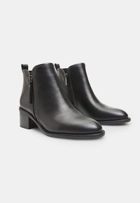 Botines de cuero negro hasta el tobillo con punta afilada, cremalleras laterales y un tacón bajo en bloque. Textura suave y acentos de metal mínimos.