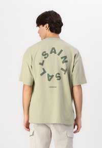 Khaki didelis medvilnės marškinėliai suapvalinta tamsiai žalia logotipo dizaino iš užpakalio, trumpomis rankovėmis ir laisvu siluetu.