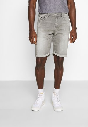 Homme portant un short en denim gris clair retroussé, des chaussettes blanches, des baskets montantes blanches et un t-shirt gris, debout sur un fond uni.