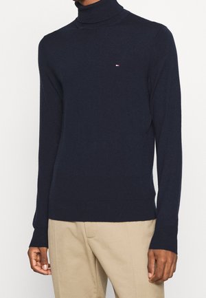 Pull en col roulé bleu marine en tissu tricoté doux avec des manches longues. Présente un petit logo sur la poitrine et des poignets et un ourlet côtelés.