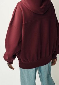 Bordeauxfarbener, übergroßer Hoodie mit Kapuze, geripptem Saum und tiefen Schultern, kombiniert mit hellblauen, weiten Jeans. Weicher Stoff, lässige Passform.