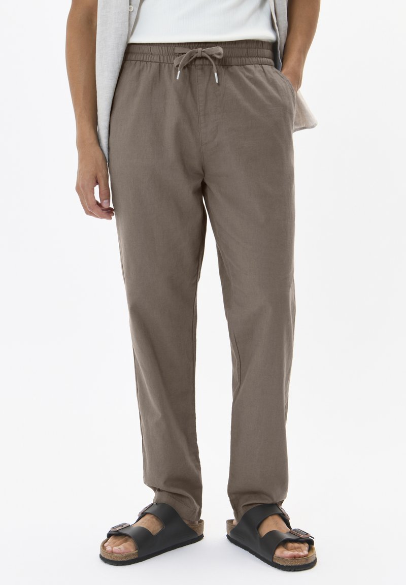 Braune Leinenhose mit elastischem Bund und Kordelzug. Kombiniert mit schwarzen Pantoletten mit Korkfußbett und verstellbarem Riemen.