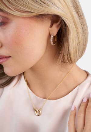 Guld hoops øreringe prydet med små glitrende sten. En delikat guld halskæde med sammenflettede ringe ses også. Blødt, lyse farvet top.