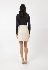 Sort tætsiddende langærmet top parret med en beige miniskirt. Modellen har lyse hæle på og står mod en ensfarvet hvid baggrund.