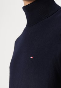 Navyfarbener Rollkragenpullover aus einem weichen Strickstoff, der ein kleines Logo mit roten, weißen und blauen Akzenten im linken Brustbereich zeigt.