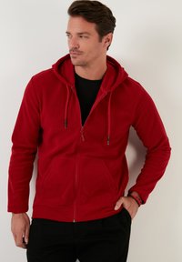 Crvena zip-up hoodie napravljena od mekog materijala, sa kapuljačom sa pertlama, prednjim dzepovima i blago prilagođenim oblikom.