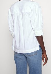 Blouse blanche à rayures avec manches bouffantes, encolure arrondie et poignets boutonnés. Fabriquée en tissu léger avec un détail plissé à l'arrière.