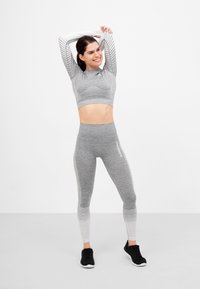 Sivi sportski crop top s rebrastim dizajnom, uparen sa sivim ombre tajicama. Crne sportske cipele. Pozadina je jednostavno bijela.