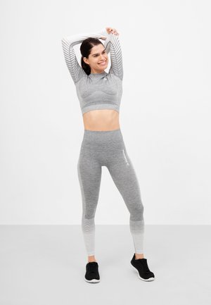 Šedý sportovní crop top s ribbed designem, doplněný šedými ombre legínami. Černé sportovní boty. Pozadí je čistě bílé.