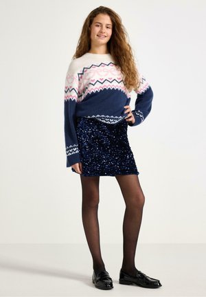Maglione lavorato a maglia con motivi geometrici rosa e blu navy, abbinato a una mini gonna blu navy con paillettes, collant neri e mocassini neri lucidi.