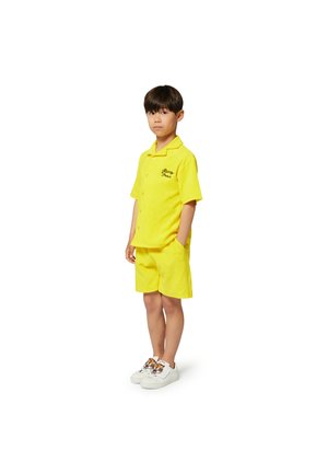 Ensemble comprenant une chemise jaune à manches courtes et un short en tissu doux, doté d'une poche et de détails brodés, associé à des baskets blanches.