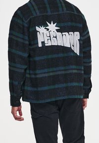 Czarna koszula flanelowa w kratę z zielonymi i niebieskimi paskami, z dużym białym logo "PEGADOR" na plecach i mankietami na guziki.