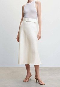 Nainen, jolla on yllään valkoinen ilmanhihoja ribbipaita, joka on työnnetty korkeavyötäröiseen kermanväriseen midi-hameeseen, jossa on leveä vyö ja vaaleanruskeat korot.