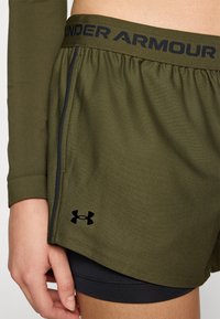 Under Armour grønne shorts laget av lett stoff, med elastisk midjebånd, svart logo, og et lagdelt design over mørke shorts.