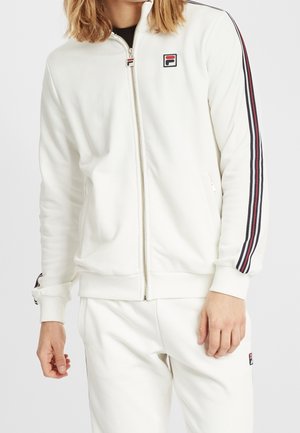 Personne portant une veste blanche zippée avec des manches rayées noires et rouges et un pantalon blanc assorti, les deux avec un petit logo.