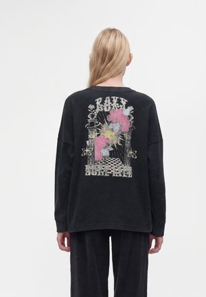 Person med langt blond hår iført en sort sweater med pink blomst og søjlegrafik samt ordene "Roxy Surf Club" på ryggen.