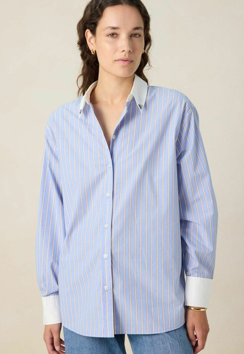 Chemise bleu clair avec des rayures verticales roses et blanches. Elle présente un col blanc, des manches longues et une coupe décontractée. Fermeture à boutons sur le devant.