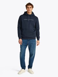 Man staande in een marineblauwe hoodie met het logo "TOMMY JEANS", blauwe spijkerbroek en witte sneakers tegen een effen witte achtergrond.