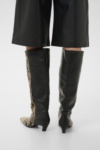Μπότες μέχρι το γόνατο με πίσω πάνελ από μαύρο δέρμα και μπροστινά μέρη με λεπτομέρεια σε σχέδιο φιδιού, φορεμένες με μαύρο παντελόνι κομμένο πάνω από τον αστράγαλο.