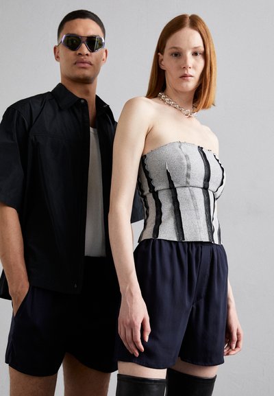 Cuplu stând împreună; femeie într-un top gri fără bretele cu linii negre, pantaloni scurți negri; bărbat în cămașă neagră, tricou alb, pantaloni scurți negri.
