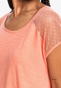 Camiseta de color rosa claro con cuello redondo, que presenta mangas de malla texturizada y un corte holgado, ofreciendo un aspecto de tejido suave y transpirable.