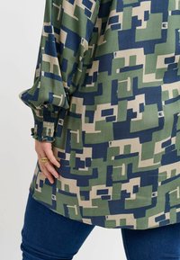 Langarmoberteil mit geometrischem Muster in Grün, Marineblau und Beige. Der Stoff ist glatt, mit gerafften Ärmelbündchen und einem lockeren Schnitt.