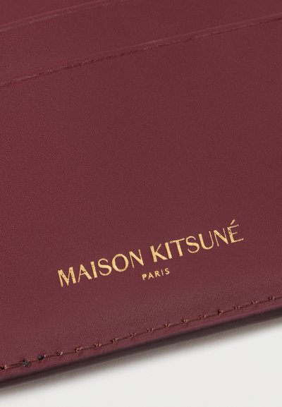 Μπορντώ δερμάτινο προϊόν με λεία επιφάνεια, διαθέτοντας ανάγλυφο χρυσό κείμενο «MAISON KITSUNÉ PARIS» και ελάχιστες λεπτομέρειες ραφής.