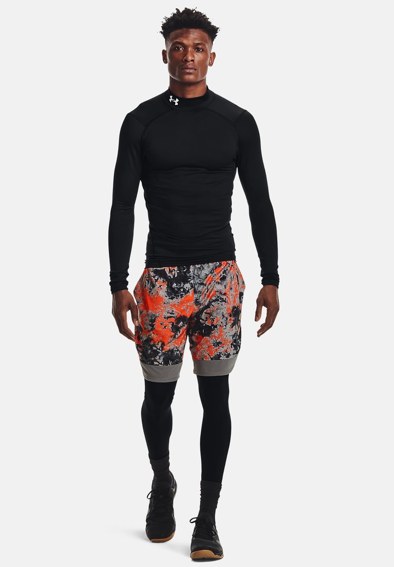 Haut de sport noir à manches longues associé à un short multicolore avec des motifs abstraits orange, gris et noir. Porté avec des chaussettes et des chaussures noires.
