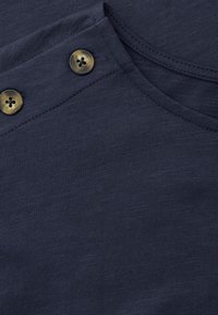 Zbliżenie granatowego materiału z dwoma brązowymi guzikami w wzór żółwia i szczegółowym przeszyciem w pobliżu zakrzywionego dekoltu.