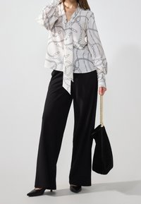 Blusa bianca con motivo a catena nera, maniche lunghe e nodo frontale, abbinata a pantaloni neri a gamba larga e a una borsa a spalla nera.