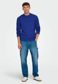 Blå kabelstrikket sweater med rund hals, parret med lyseblå jeans og hvide sneakers. Sweateren har ribbede manchetter og kant.