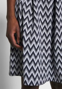 Materiał w czarno-białym wzorze w zygzaki z plisowanym wykończeniem. Zbliżenie ręki opierającej się na krawędzi, podkreślające teksturę i szczegóły wzoru.