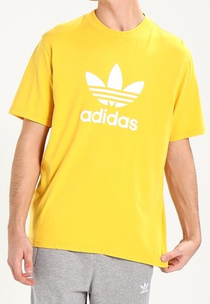 Žluté bavlněné tričko s velkým bílým logem Adidas a designem tří pruhů na přední straně. Volný střih s kulatým výstřihem.