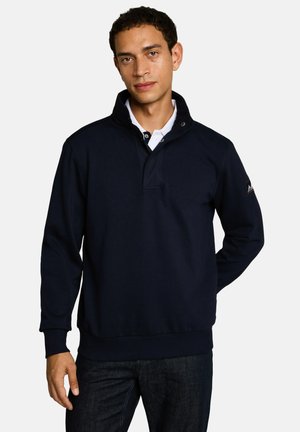 Mann trägt einen marineblauen Pullover mit Stehkragen über einem weißen Hemd mit Kragen und steht mit einer Hand hinter dem Rücken.