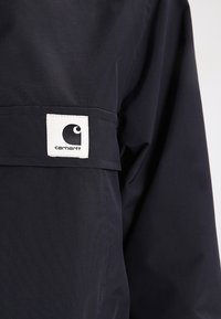 Navy blue jacket made of durable fabric features a white square patch with a black logo. Smooth texture with a simple design.