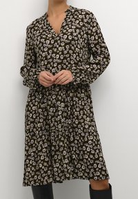 Femme portant une robe noire à manches longues ornée de petites fleurs blanches et violettes, se tenant les mains jointes, devant un fond clair.